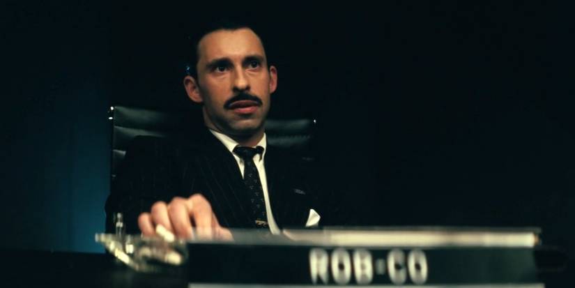 Рафі Сілвер у ролі Роберта Хауса, сидячи перед табличкою з назвою Rob-Co у серіалі "Фолаут"